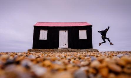A woman leaps from the side of a small black and red hut on the riverbank in Rye Harbour Nature Reserve, East Sussex UK